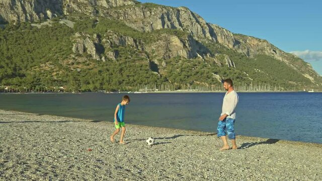 Young bearded man and his son playing football by the sea. Young father having fun and spending time together with his little son at the sea view. Happy family enjoying vacation.