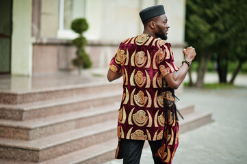 Back of african stylish and handsome man in traditional outfit and black cap standing outdoor.