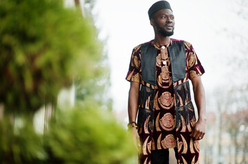 African stylish and handsome man in traditional outfit and black cap standing outdoor.
