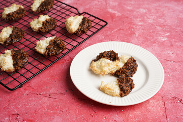 Several coconut flakes cookies dipped in chocolate on a white plate and baking rack on magenta surface, top view
