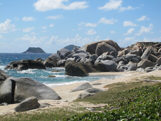 A beach of boulders, stones and rocks with green vegetation and shiny blue sea. Green island in the background. Virgin Gorda, British Virgin Islands, Caribbean.