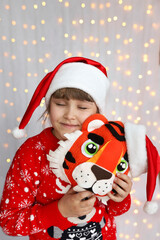 Close-up portrait of a cute dreamy Christmas girl in red Santa hat and in a red Christmas sweater with closed her eyes. Child with hand made toy tiger. Background of defocused lights.