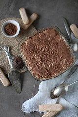 Tiramisu cake in a crystal square large plate on the table with a spruce towel and spoons. Dessert with mascarpone and savoyardi cookies.