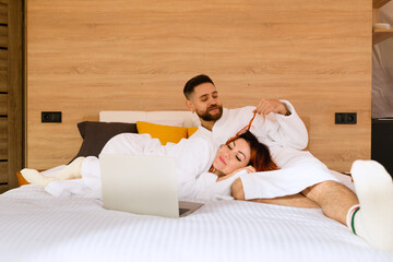 A young couple in bathrobes is lying on the bed with a laptop. A man playing with a woman's hair. A woman lies with her head in the lap of a man.