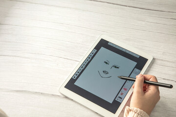 Young creative designer holding stylus pen drawing on screen of digital tablet on wooden desk with copy space, modern digital art top view