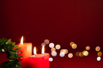 Scena natalizia con tre candele rosse accese e una ghirlanda di vischio, con luci sfocate su sfondo rosso con spazio per scrivere. Natale. Auguri. Dicembre. Inverno. Feste. Feste invernali.
