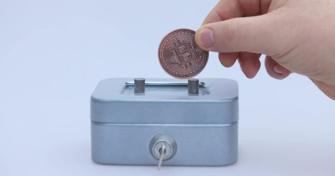Male hand puts a cryptocoin, Bitcoin or BTC, in a small grey vault, safe or piggy bank, depicting crypto or cryptocurrency saving, holding or hodling. High quality 4k footage