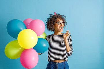 Cheerful multiracial lady in birthday cone hat looking away and laughing while holding multicolored balloons. Isolated on blue background
