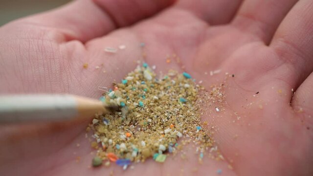 Global ocean pollution concept with microplastics bits and pieces from single-use plastic consumer items. Selective focus of a person examining micro plastics in his hand with a pencil.
