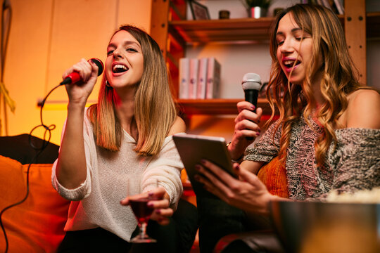 Two female friends sit at home on Saturday night and sing a duet on karaoke night.
