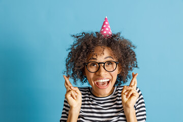 Cheerful multiethnic lady in birthday cone hat doing fingers crossed gesture and laughing while making wish. Isolated on blue background