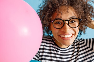 Close up of joyful lady in glasses looking at camera and smiling while standing near birthday balloon. Isolated on blue background