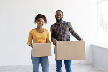 Smiling millennial african american woman and man carry cardboard boxes with things in empty room