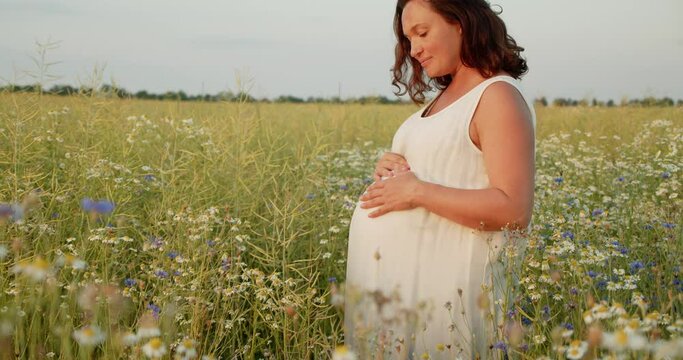 Pregnant woman with a big belly feeling happy outdoors. Adult expecting mother stroking belly on nature.  Middle age white pregnant woman touching her belly while sunset. Woman pregnancy concept.