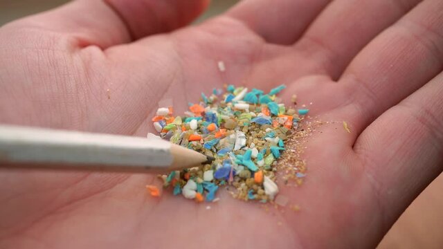 Global ocean pollution concept with microplastics bits and pieces from single-use plastic consumer items. Selective focus of a person examining micro plastics in his hand with a pencil.