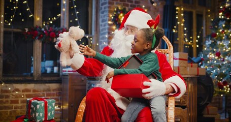 Little multiracial girl sitting at the Santa knees and taking off her gift from the present box with interest. Santa Claus looking at her with smile