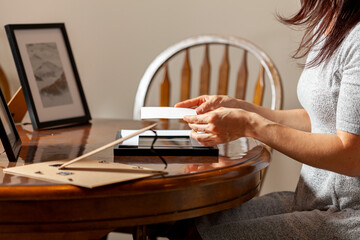 A young caucasian woman is placing a printed photograph into a picture frame with kickstand. There are other frames on the table. Tabletop picture framing concept.