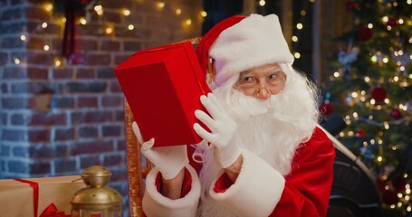 What is inside. Bearded Santa Claus shaking with big red present box and guess what inside while spending time at the room with Christmas decorations at the background. Holidays spirit concept