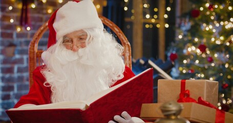 Fototapeta na wymiar Santa Claus reading book while sitting at the armchair with presents with New Year decorations at the background. Merry Christmas concept