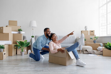 Fototapeta na wymiar Happy black man pushing laughing wife in carton box. Energetic woman sit in cardboard container, have fun