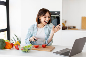 Prepare food at home. Positive young lady cutting vegetables and preparing salad, talking on video chat via laptop
