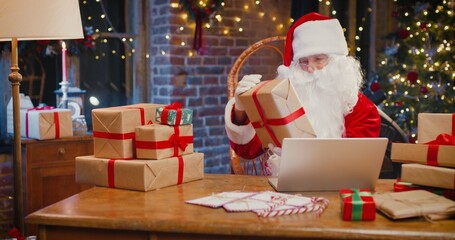 Close up of happy senior man in Santa Clause costume checking presents with the laptop while sitting at home. Holidays concept