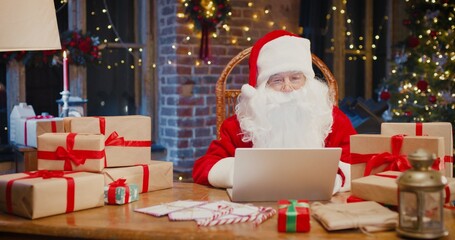 Cheerful white-haired Santa Claus sitting at the armchair and working at the laptop with many Christmas presents and gift boxes around. Holidays spirit concept