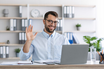 Obraz premium Smiling mature european guy with beard in glasses waving hand to laptop webcam in office or home interior