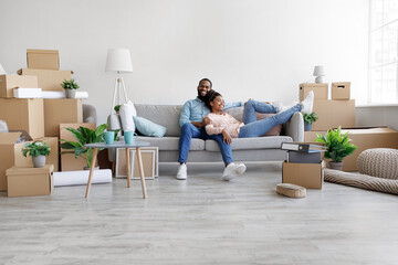 Happy glad young african american husband and wife relaxing on couch in living room with cardboard boxes