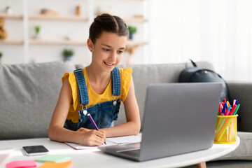 Cheerful teen sitting at desk, using laptop, drawing in copybook