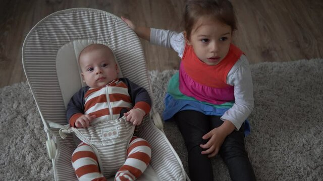 Childhood, motherhood, family, comfort. Little newborn baby boy in striped bodysuit pajamas awake looking around smiles plays rattles sibling sister kid lul amuse brother in room at home atmosphere