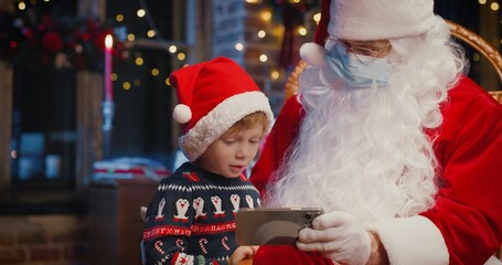 Fototapeta na wymiar Little caucasian boy using smartphone with Santa Claus wearing protective mask while spending time at home with New Year decorations. Pandemic concept