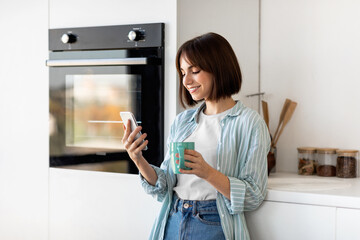 Fototapeta na wymiar Social networks and chat in morning. Happy young lady drinking coffee and using smartphone, standing in kitchen
