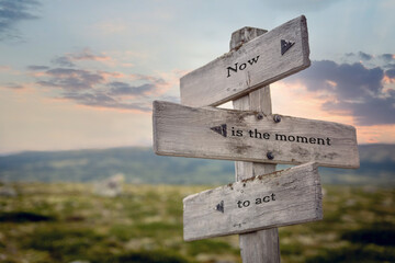 now is the moment to act text quote on wooden signpost outdoors in nature during sunset.
