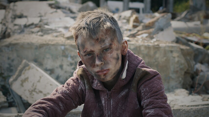 Teen survivor with dirty face and clothes looking at camera while sitting near ruins of destroyed...