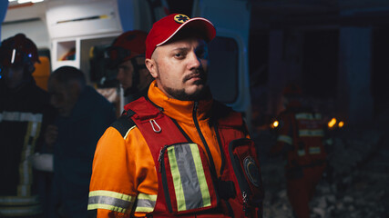 Male paramedic in uniform and hat looking at camera while standing near ambulance car during rescue mission after disaster at night