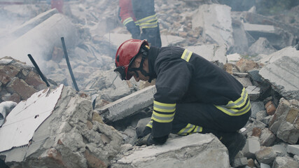 Pan left view of male emergency workers removing concrete rubble in cloud of dust while working on ruins of destroyed building after disaster - obrazy, fototapety, plakaty