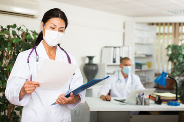 Professional female doctor wearing protective face mask standing in clinic office, carefully reading patient medical card, analyzing diagnosis