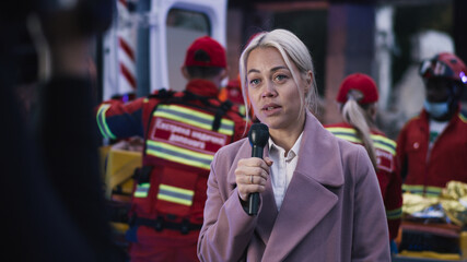 Blond woman with microphone making report near team of paramedics on disaster site after earthquake