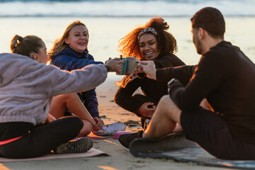 Multiethnic group of people toasting with tea sitting on the beach during sunset
