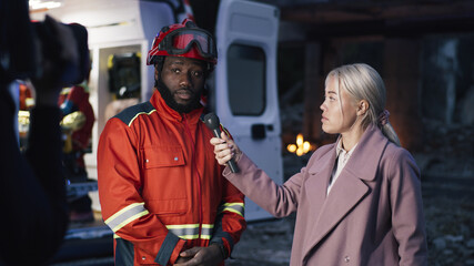 Blond woman in coat asking questions to African American paramedic near van while reporting from disaster site in evening © Framestock