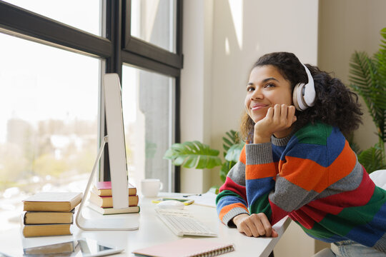 Portrait Of Pensive Beautiful African American Woman Wearing Headphones Sitting At Workplace Looking Away. Freelancer Using Computer Working From Home  