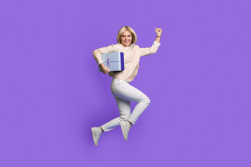 Full body photo of a happy caucasian woman jumping up holding in arm holiday gift isolated over purple color background. Holiday gift surprise.
