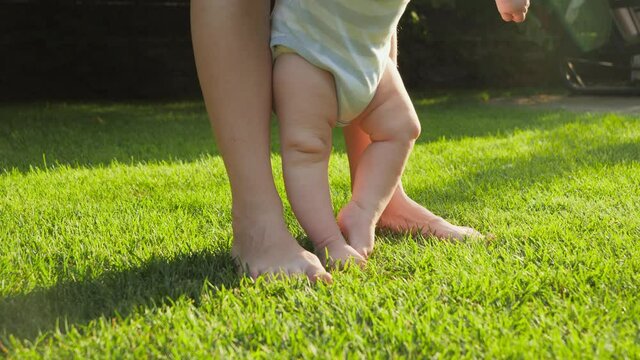 Closeup of barefoot baby with mother standing on fresh green grass lawn at house backyard garden. Concept of healthy lifestyle, child development and parenting.