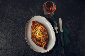 Plate with tasty Ajarian khachapuri and glass of wine on dark table background