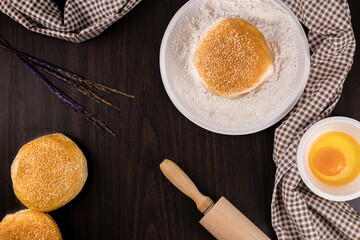 bread concept buns, a dish of flour, a rolling pin, and a tiny bowl of raw egg lying on the dark wooden table
