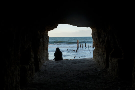 Girl sitting in a cave opening, facing the beach with jetty ruins in Port Willunga beach, Adelaide South Australia