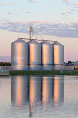 Obraz premium Row of metal grain silos with overhead conveyor reflected in adjacent water body, illustrating agricultural storage infrastructure and rural crop handling operations