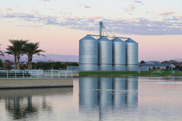 Four large metal grain silos reflected in a calm water retention pond at sunrise, illustrating agricultural infrastructure integrated within a suburban landscape © Eduardo Barraza
