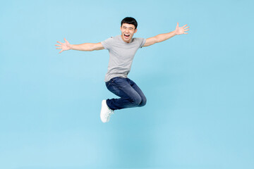 Jumping ecstatic young Asian man with arms stretching in mid air isolated in blue studio background